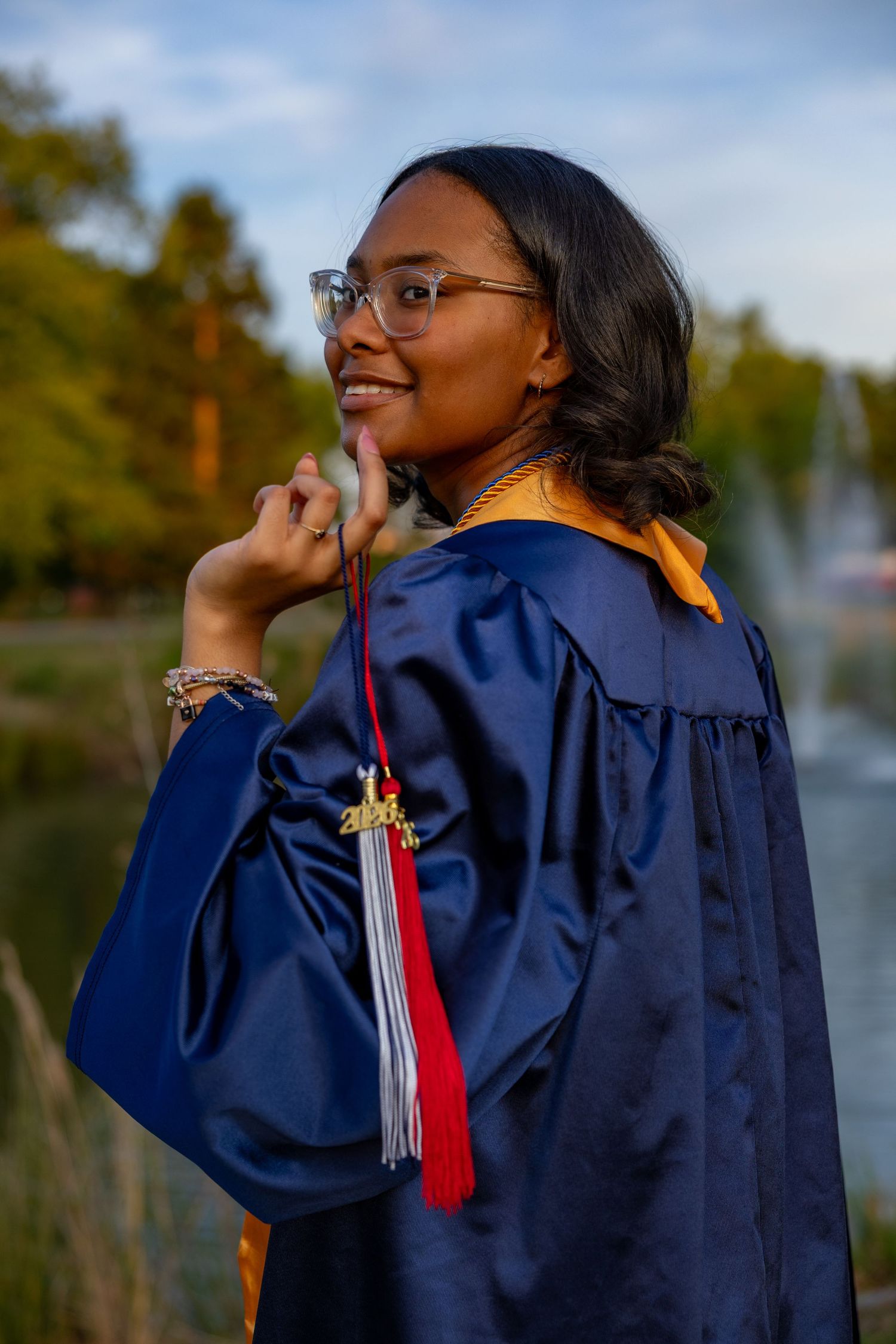 Young woman in blue graduation gown and glasses smiles proudly outdoors near a pond with trees in background.