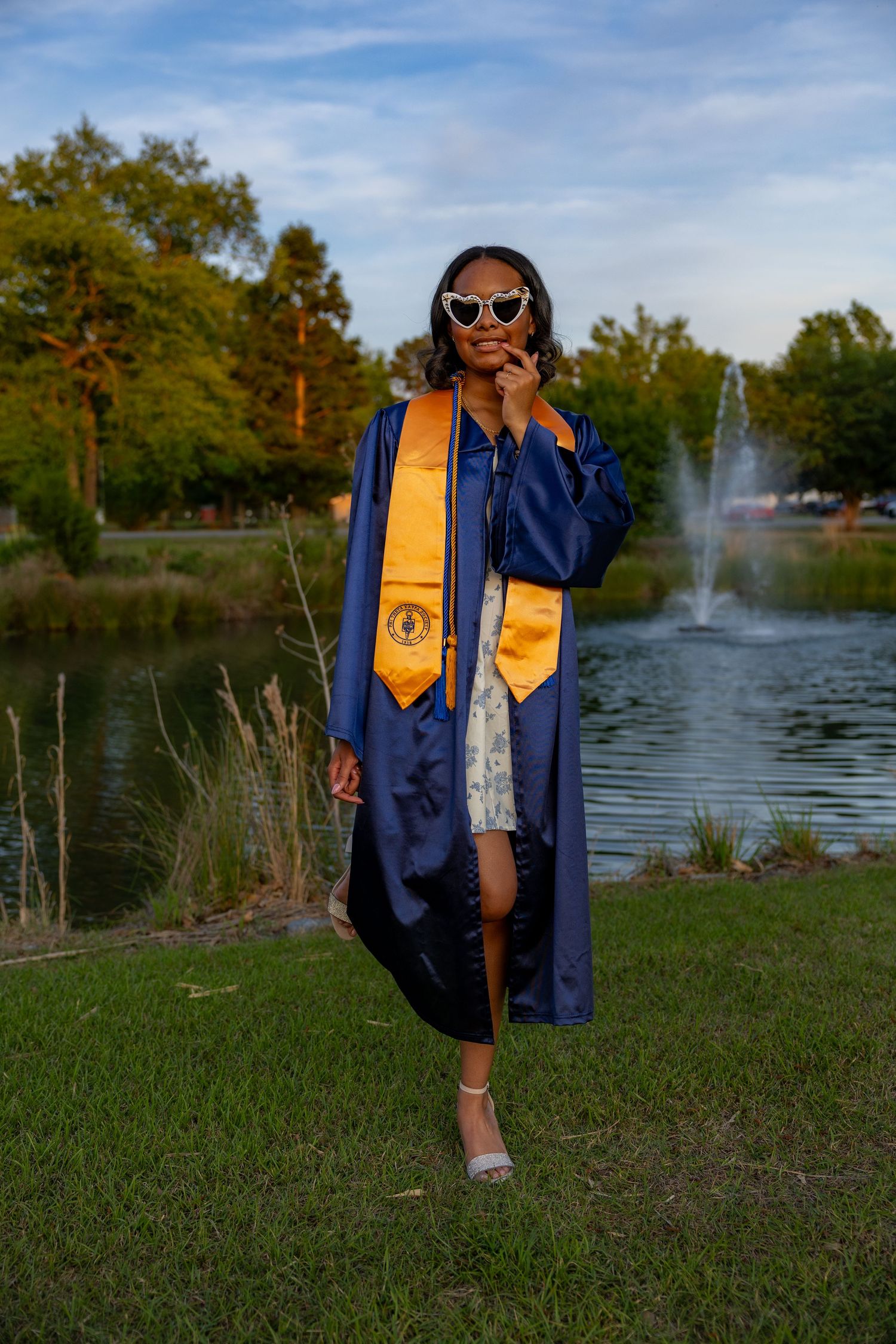 Young woman in blue graduation gown and gold stole poses by a pond with fountain, wearing white sunglasses.