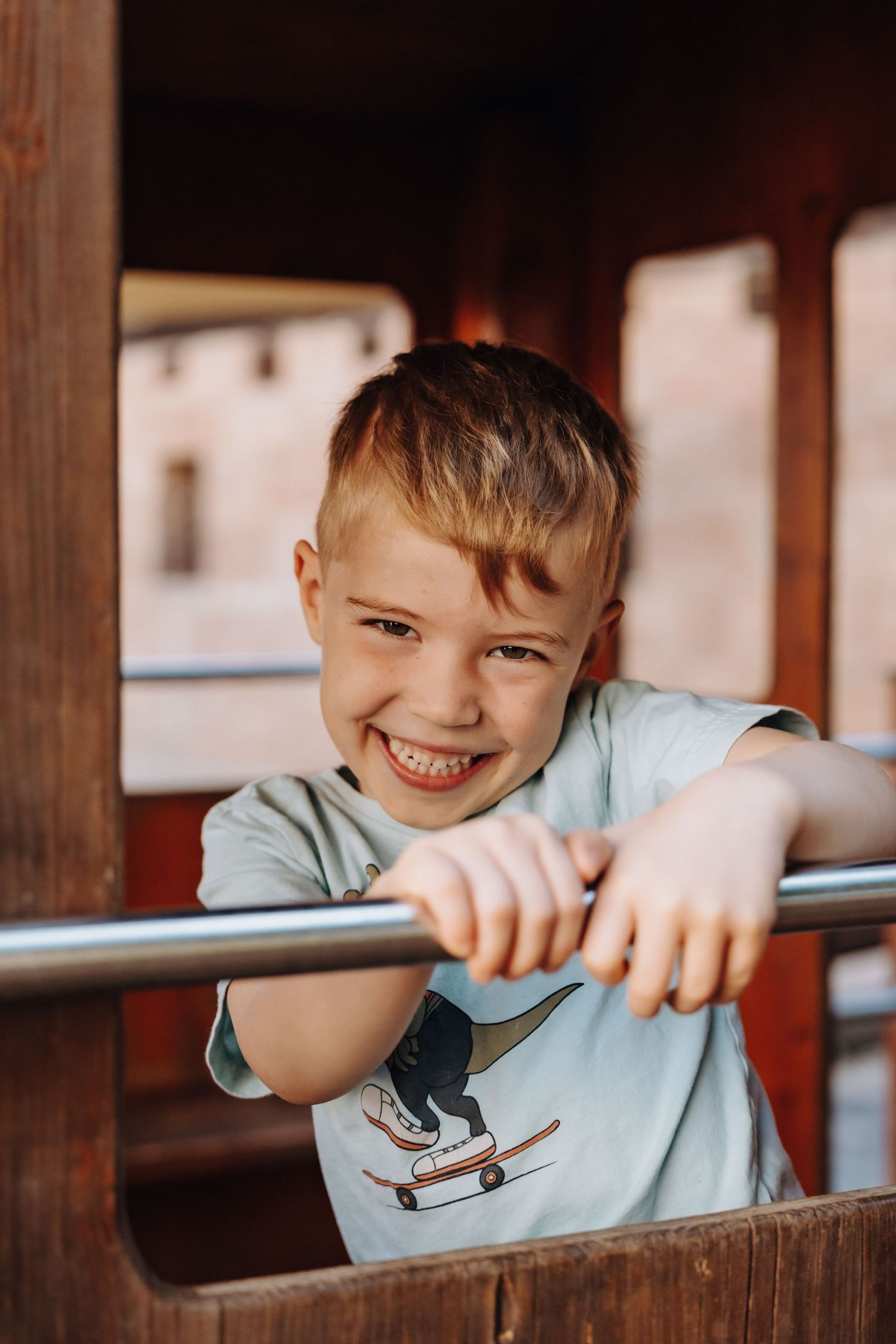 Happy young boy smiling while gripping a metal bar on a wooden playground structure outdoors.
