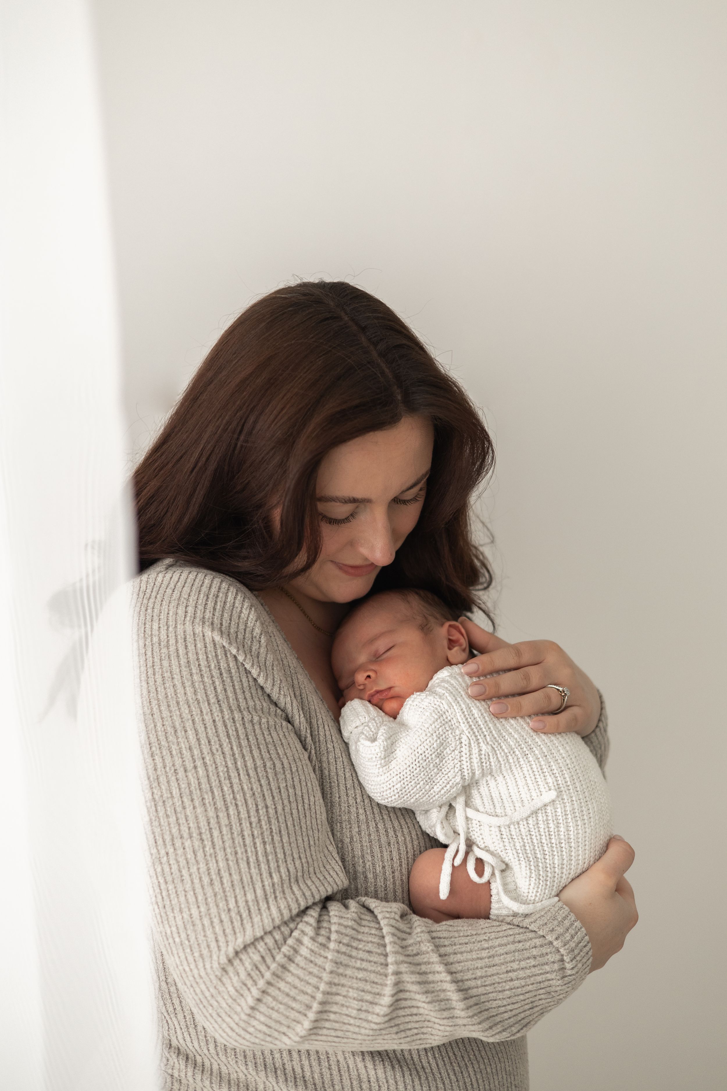 A mother tenderly holding sleeping newborn baby dressed in white knit outfit against white wall timeless Nottingham newborn photographer