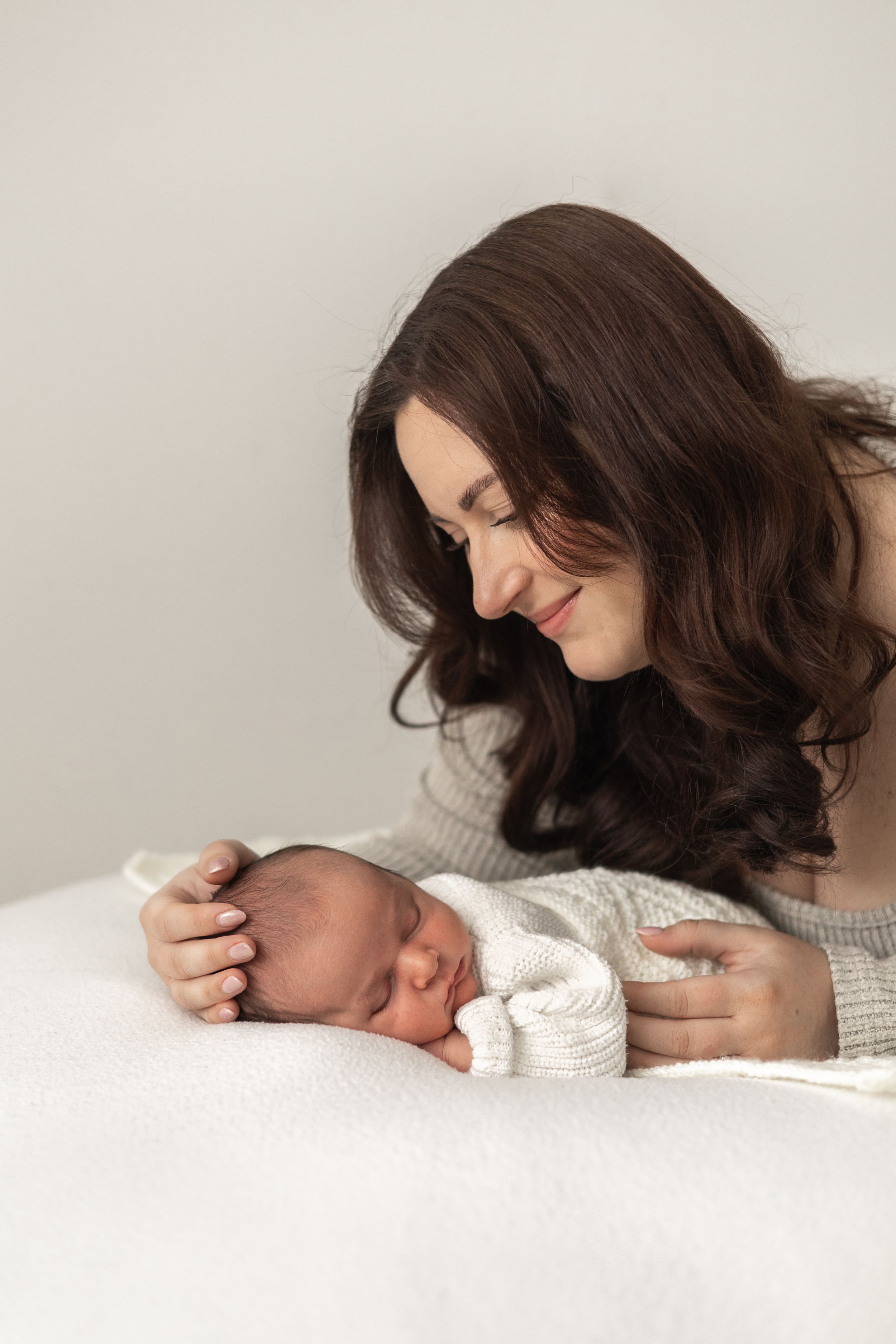 A loving mother with long brown hair smiles tenderly at her sleeping newborn baby wrapped in white. timeless Nottingham newborn photographer