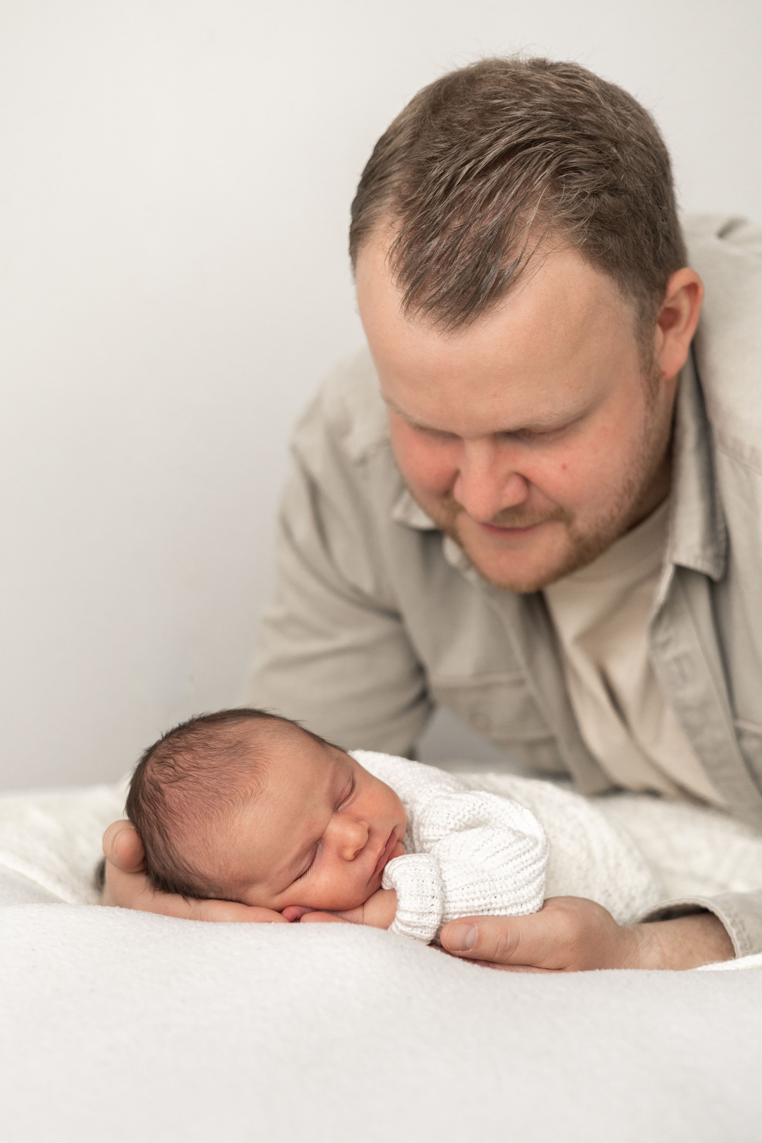 A dad tenderly gazes at his sleeping newborn baby wrapped in white, lying on a soft blanket timeless Nottingham newborn photographer