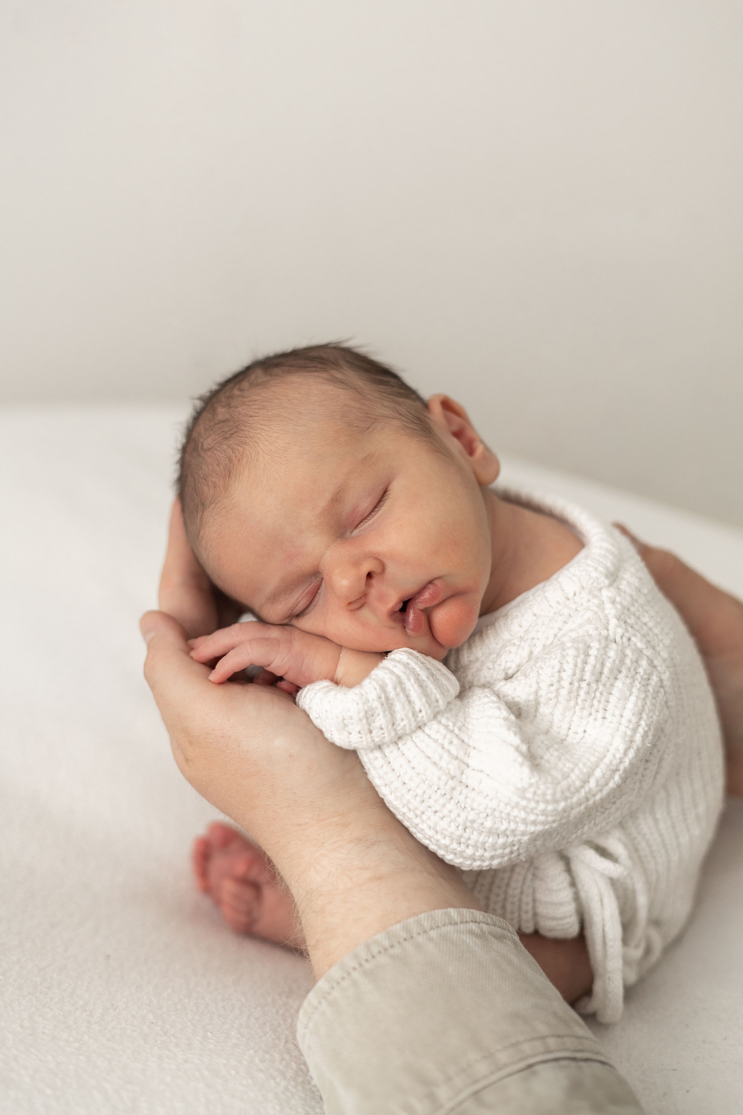 Sleeping newborn baby in white knit outfit cradled gently in dads hands on soft white surface  timeless Nottingham newborn photographer