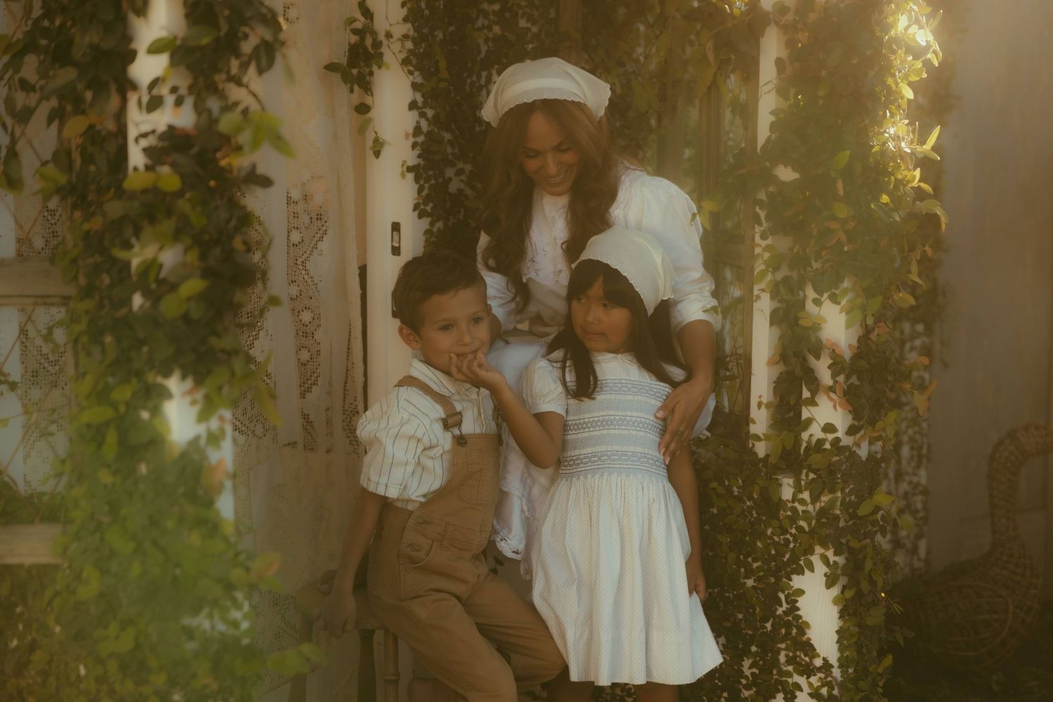 An Orange County family photographer captures a woman in a white dress with two young children outdoors amid lush green ivy backdrop.
