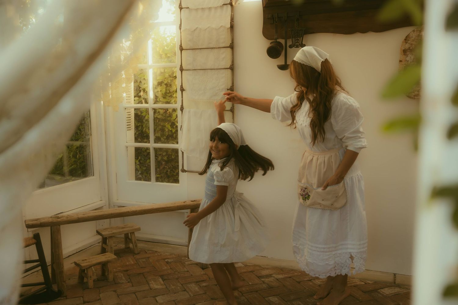 Two girls in white dresses dance together in a charming rustic room with French doors and warm natural light.