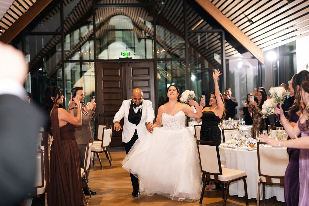 Bride and groom make grand entrance at elegant wedding reception hall, guests cheering and applauding around decorated dining tables.