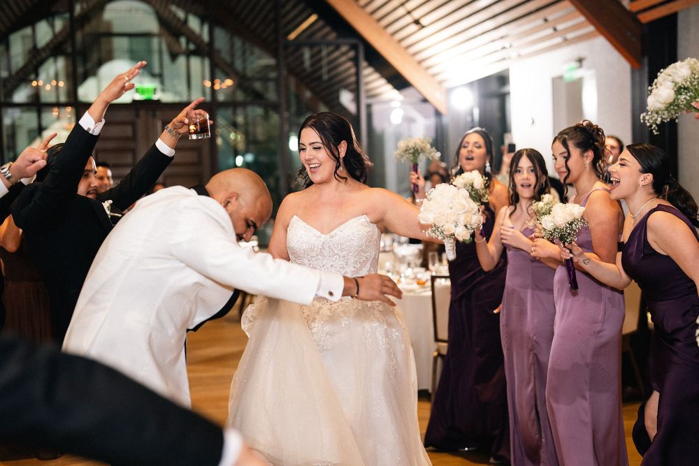 Bride and groom dancing joyfully at wedding reception surrounded by bridesmaids in purple dresses holding white bouquets.
