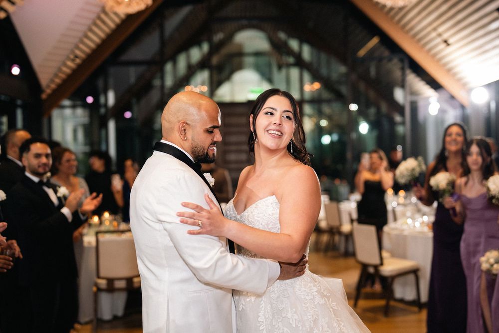 Bride and groom share their first dance at elegant wedding reception venue with guests applauding in background.