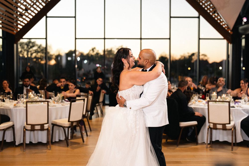Bride and groom share first dance kiss in elegant wedding venue with floor-to-ceiling windows and candlelit reception tables.