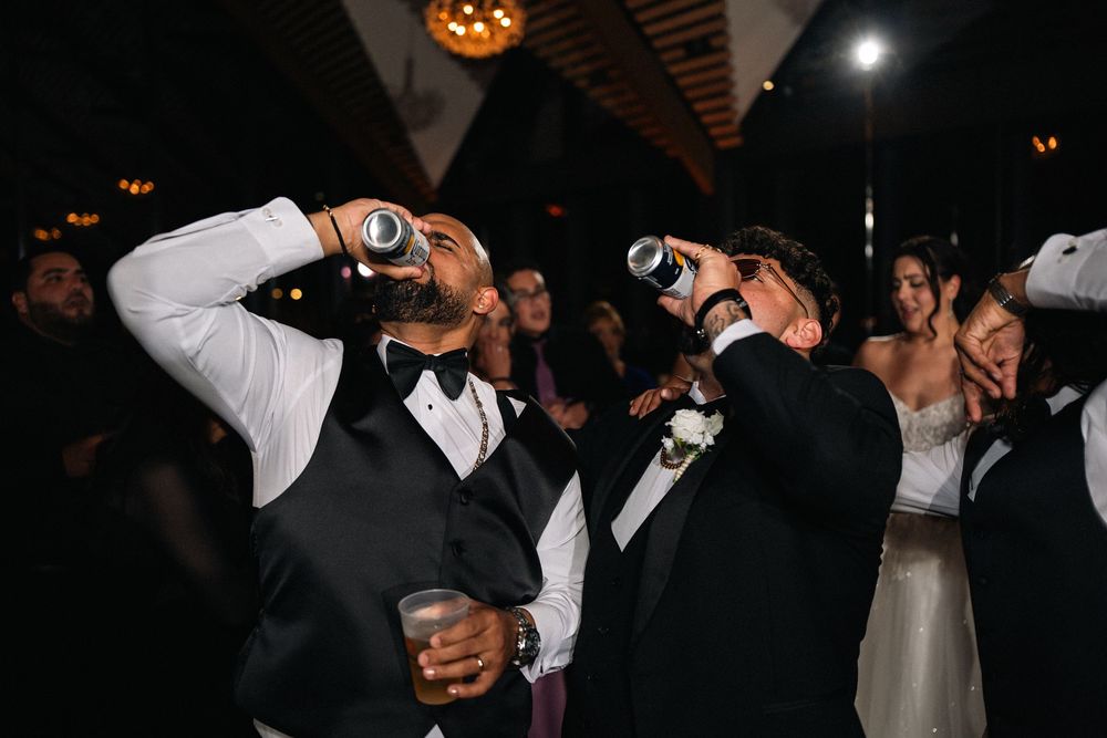 Two groomsmen drinking from bottles at a lively wedding reception celebration on the dance floor.