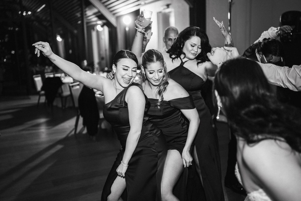 Women in black dresses dancing joyfully at a wedding reception in a black and white photo.