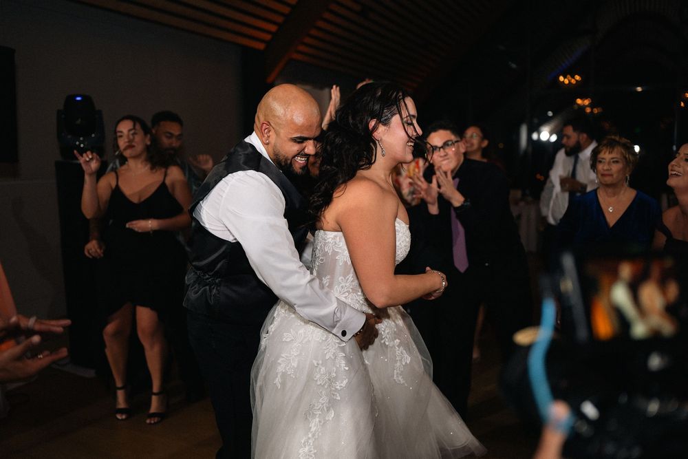 Bride in white gown and groom in vest share first dance at wedding reception with guests applauding in background.