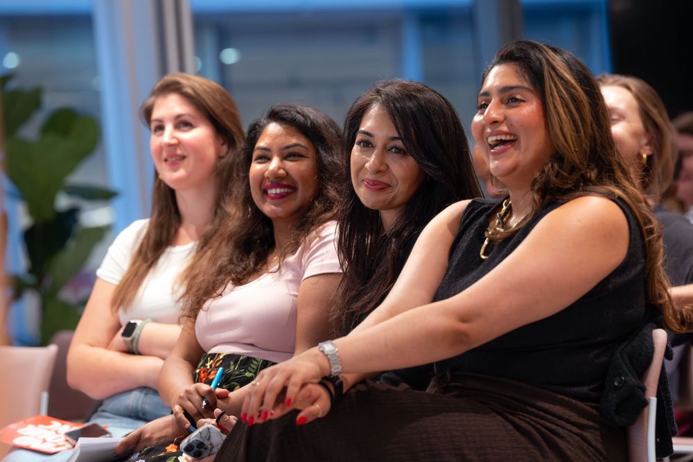 Four women laughing and smiling together at an indoor event, enjoying a presentation or performance.