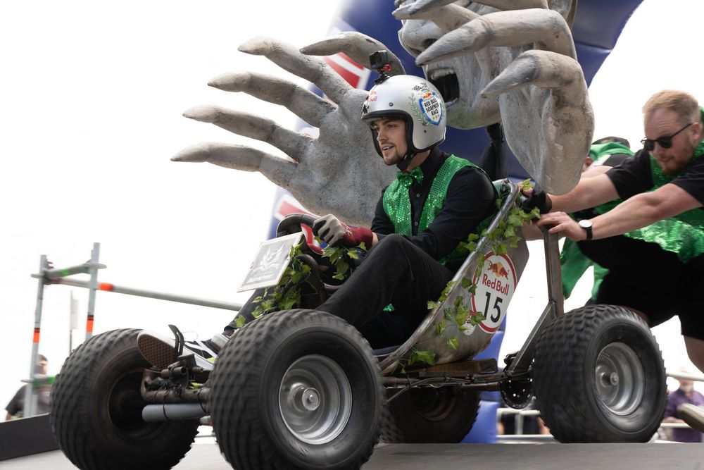 Person in helmet driving a camouflage go-kart at an outdoor racing event with a large monster sculpture behind.