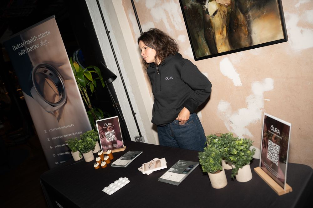 Woman standing at a display table with plants, brochures, and a banner at an indoor event booth.