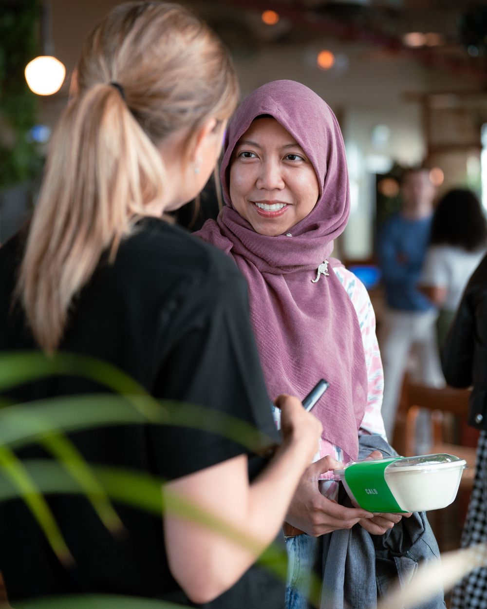 Two women conversing in a cafe, one wearing a pink hijab and holding a green notebook.