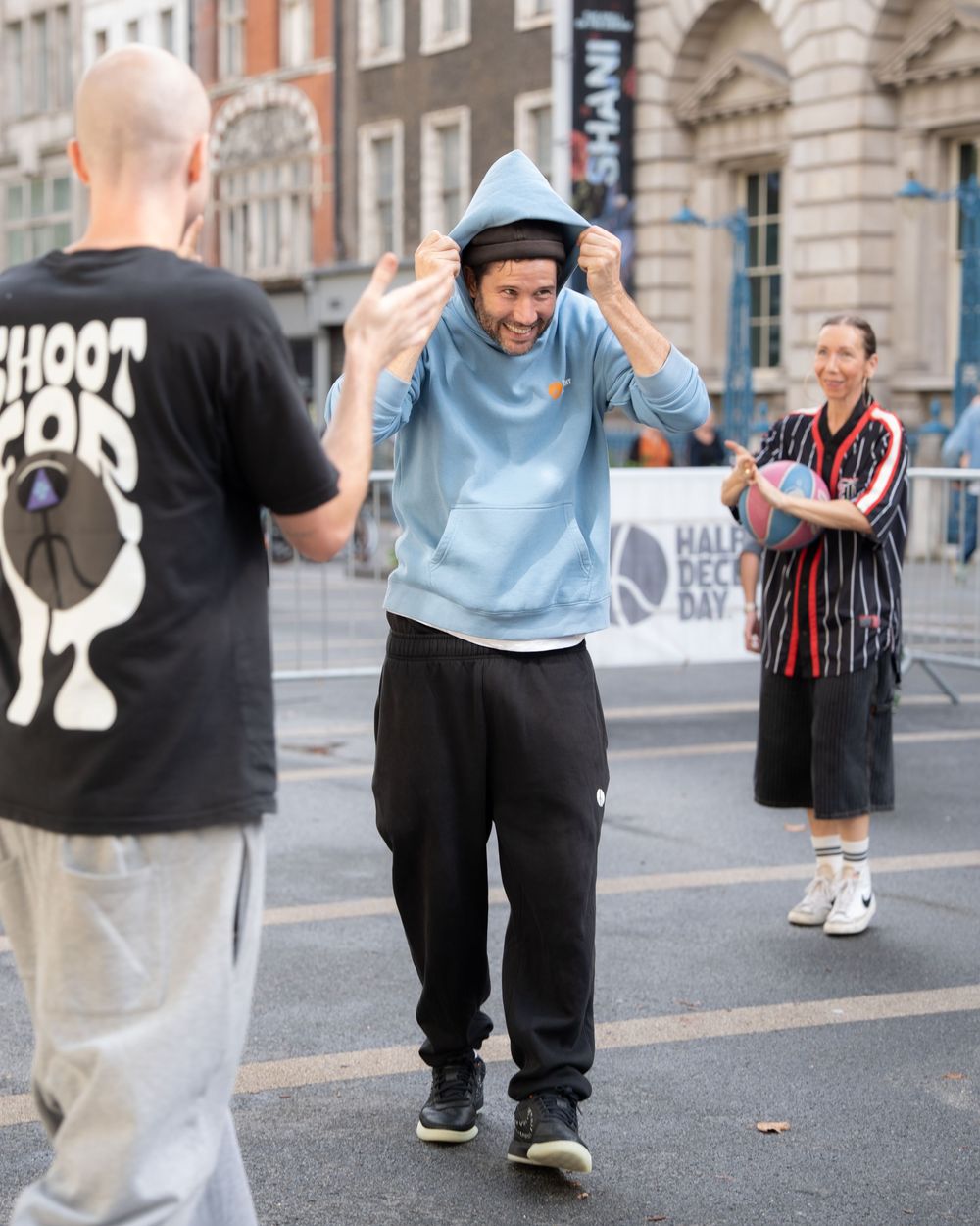 Man in blue hoodie laughing outdoors at a street event, with people in background on an urban street.