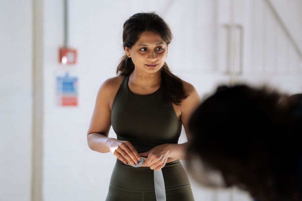 A fit woman in olive sportswear wraps her hands with boxing wraps while preparing for a training session indoors.