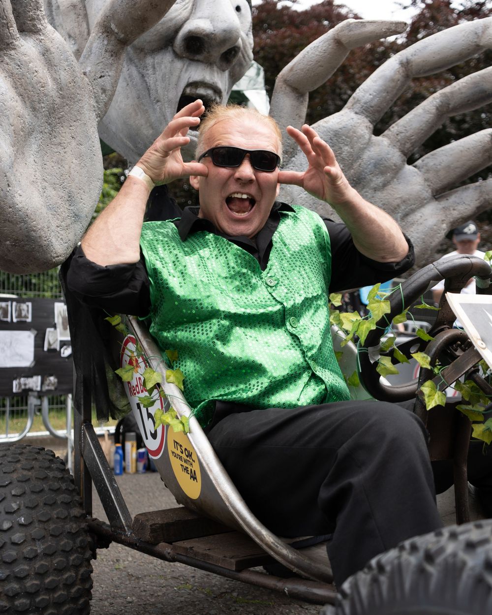 Man in green sequin vest sitting in decorative cart, making scary claws gesture in front of giant hand sculpture.