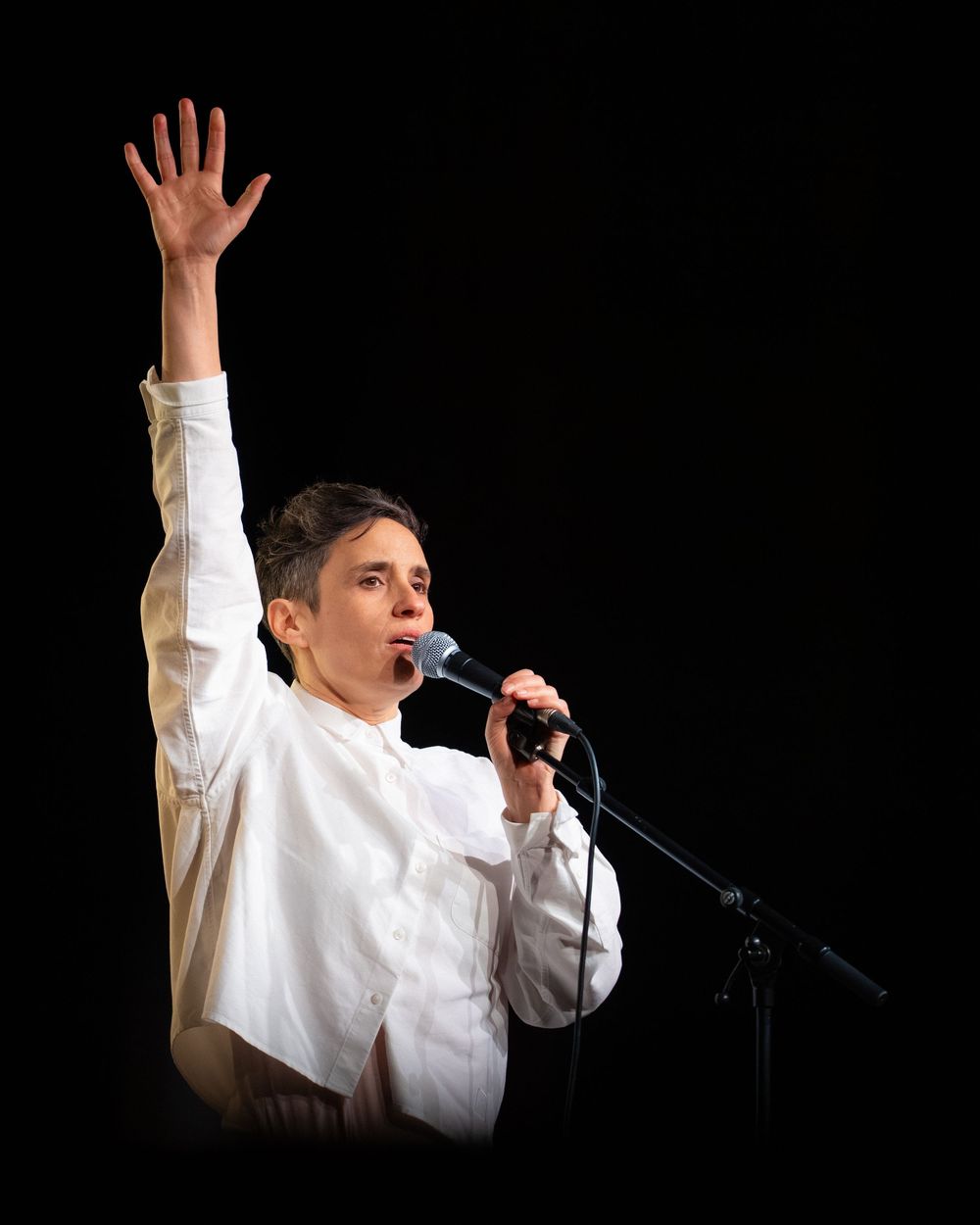 Performer in white shirt singing into microphone with one arm raised against black background.