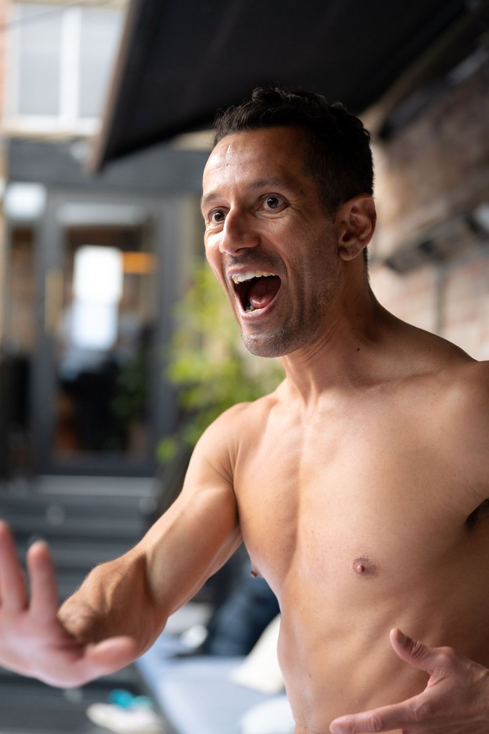 Shirtless muscular man laughing and gesturing with hand in modern indoor setting.