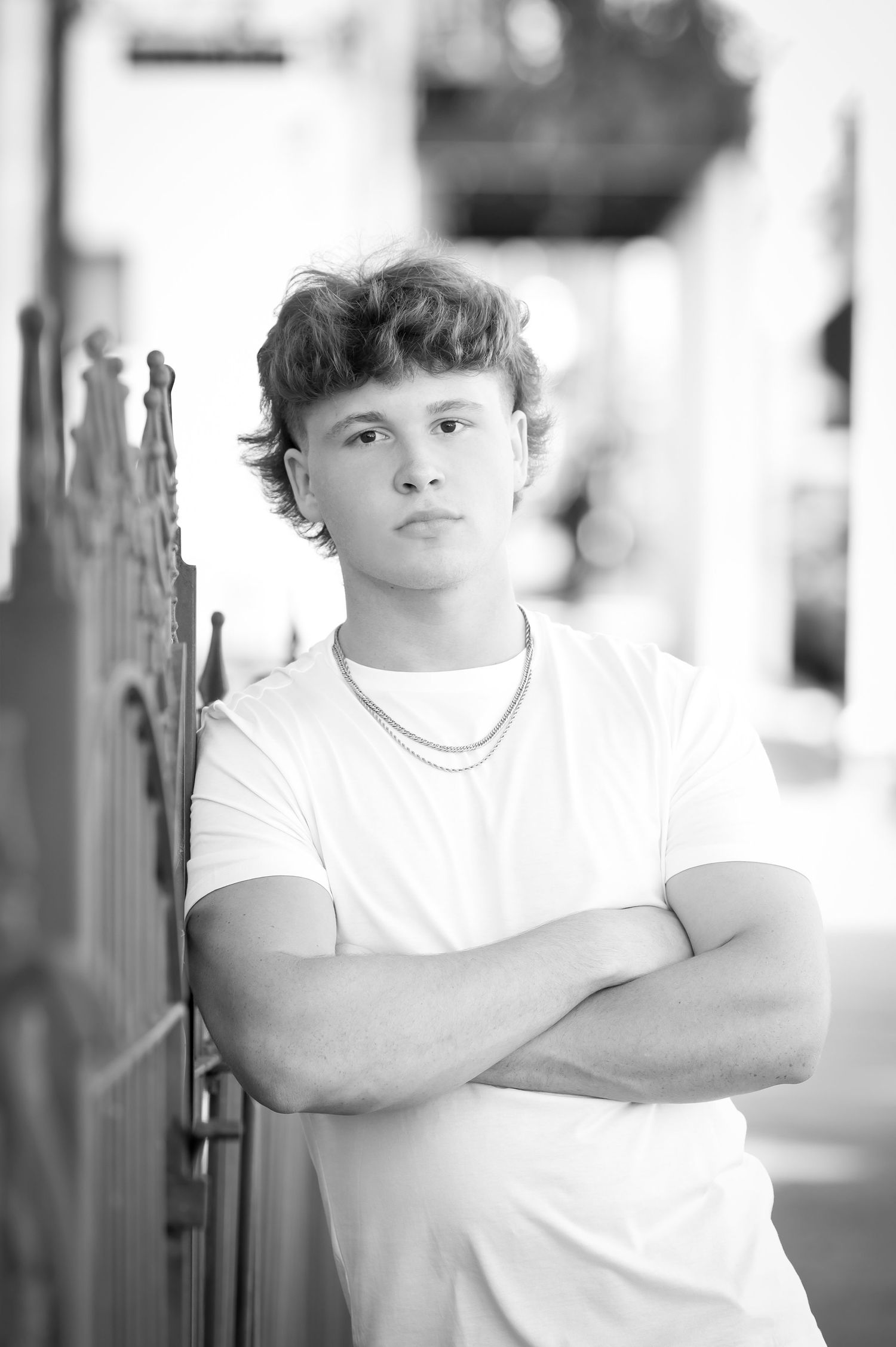 Young man with curly hair crosses arms, leaning against iron fence in black and white portrait photo.