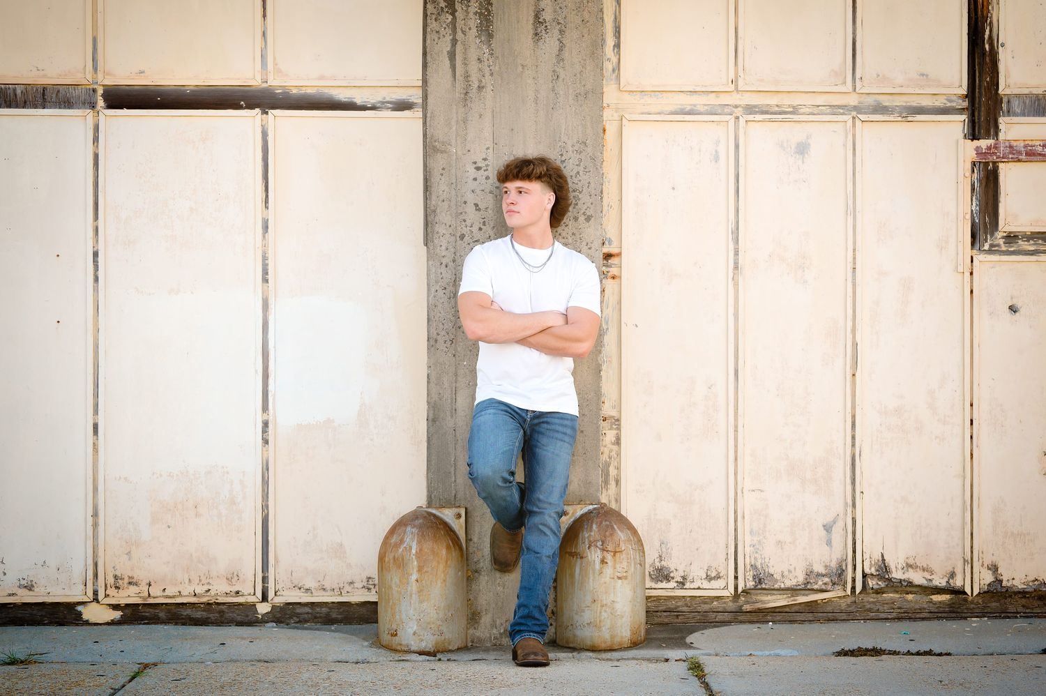 Teen boy in white polo and jeans leans against rustic wooden doors with arms crossed outdoors.