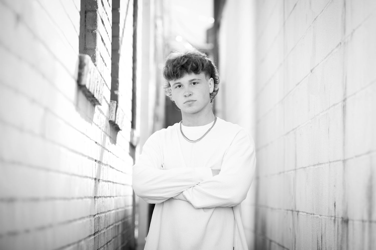 Teen boy in white sweatshirt poses arms crossed in alley for black and white senior portrait.