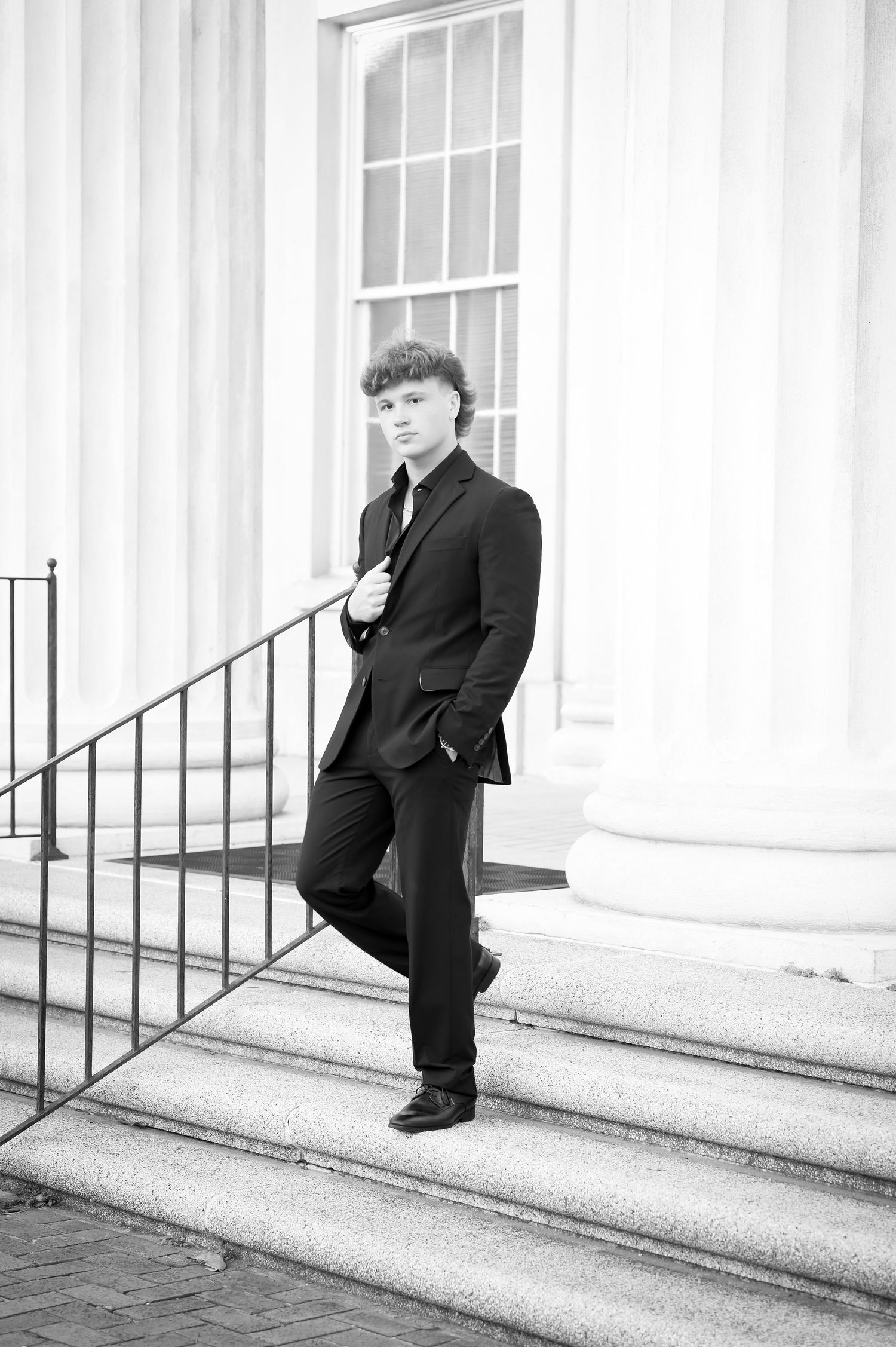 Young man in black suit posing on steps by white columns in elegant black and white senior portrait.