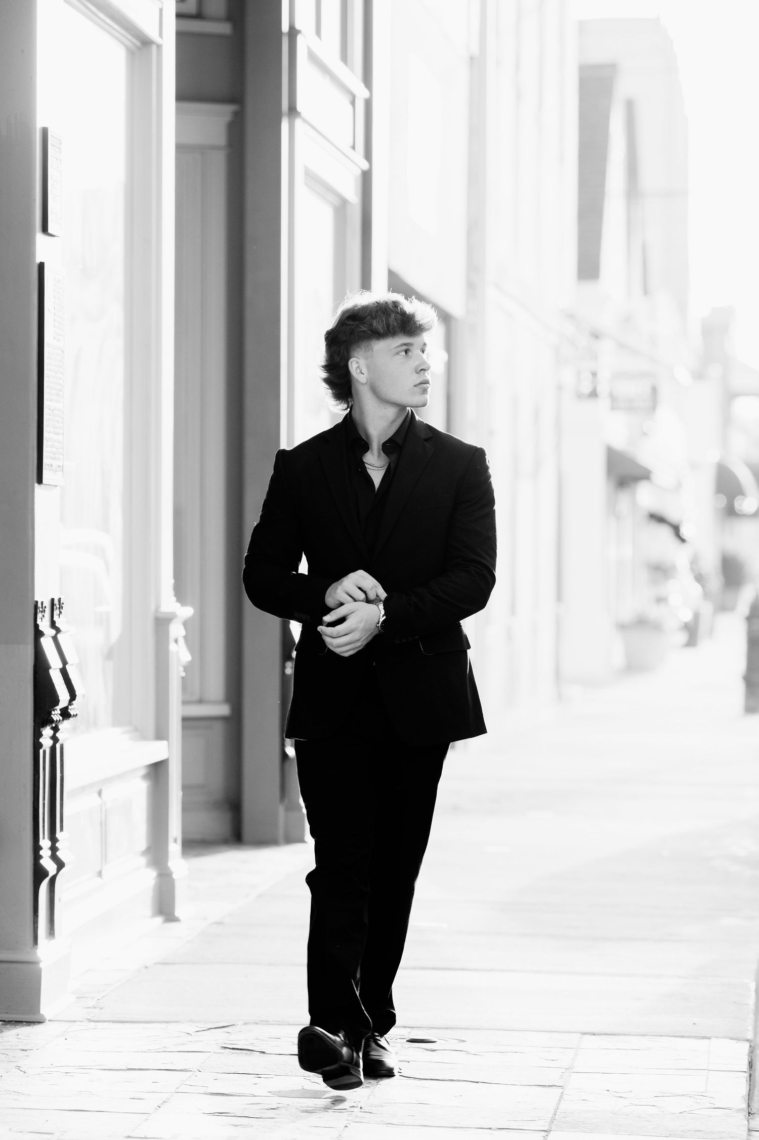 Black and white photo of a young man in a dark suit walking confidently along an urban sidewalk.