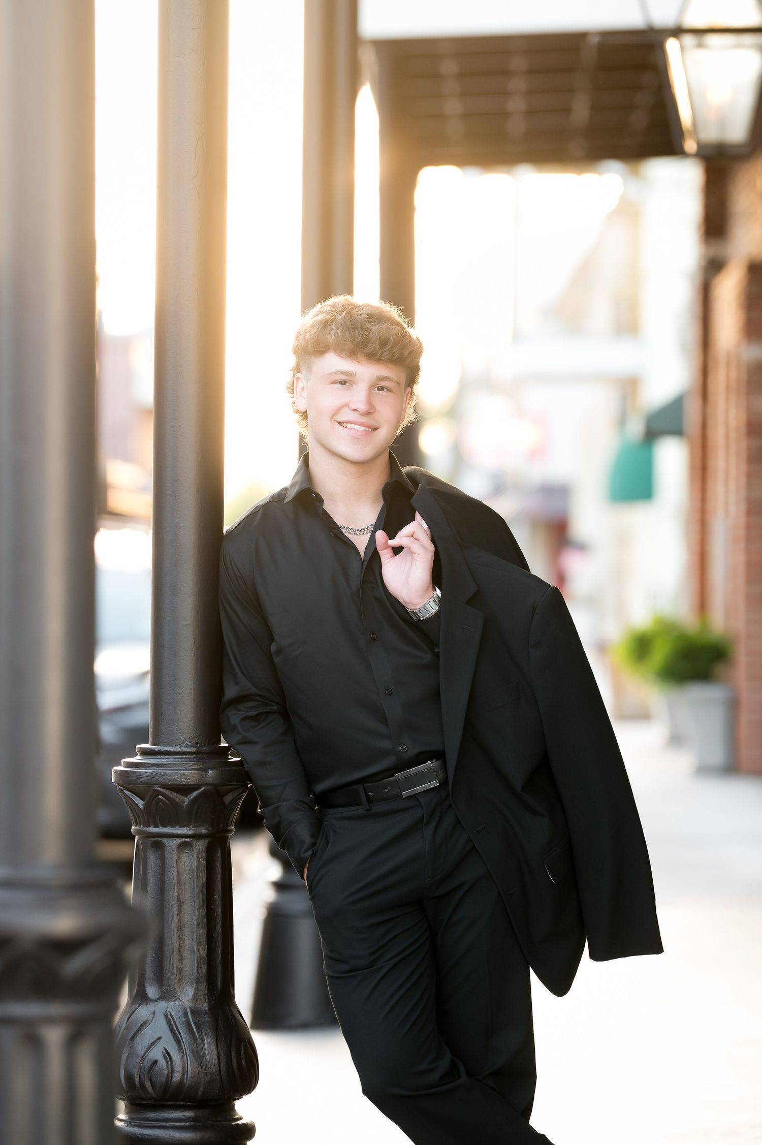 Young man in black shirt and blazer leans on lamppost smiling in golden hour urban setting.