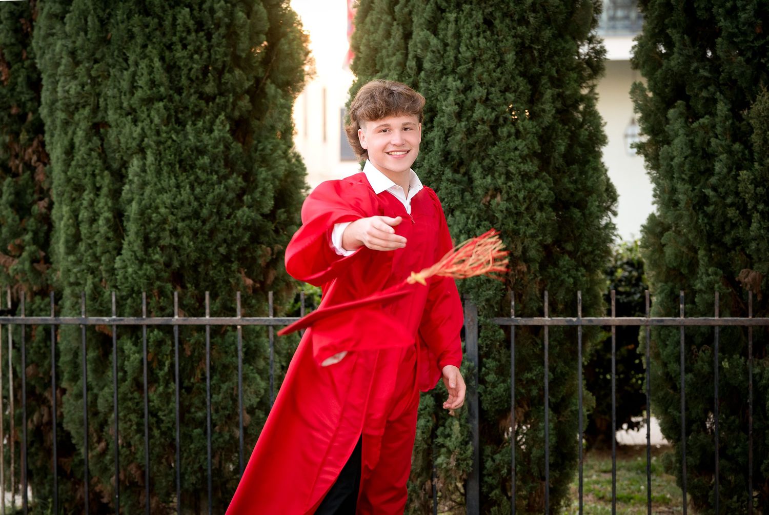 Smiling boy in red jumpsuit holds lacrosse stick near iron fence with tall cypress trees behind.