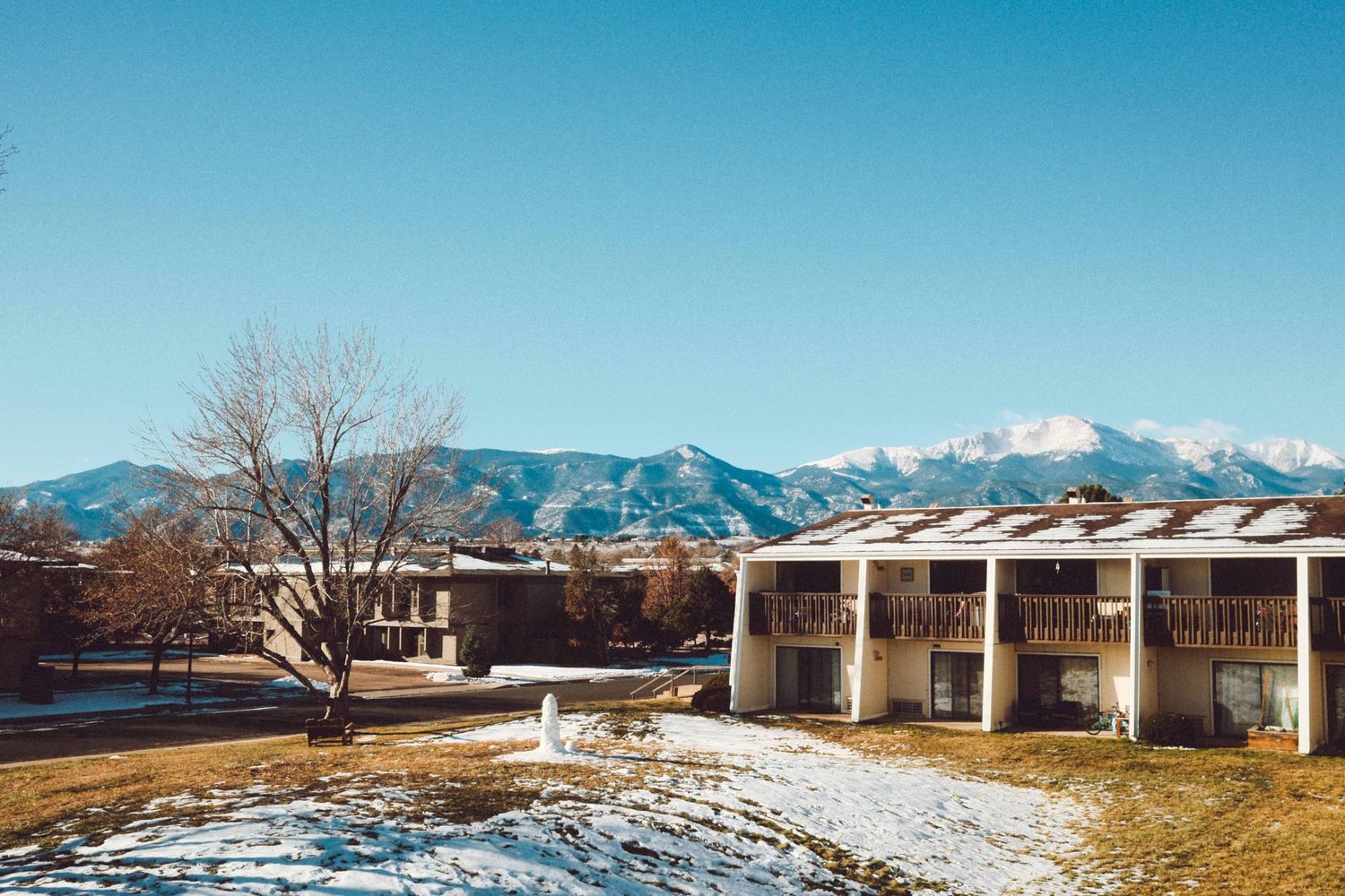 Snow-dusted mountain resort with balconied building, bare tree, and Rocky Mountains backdrop under clear blue winter sky.