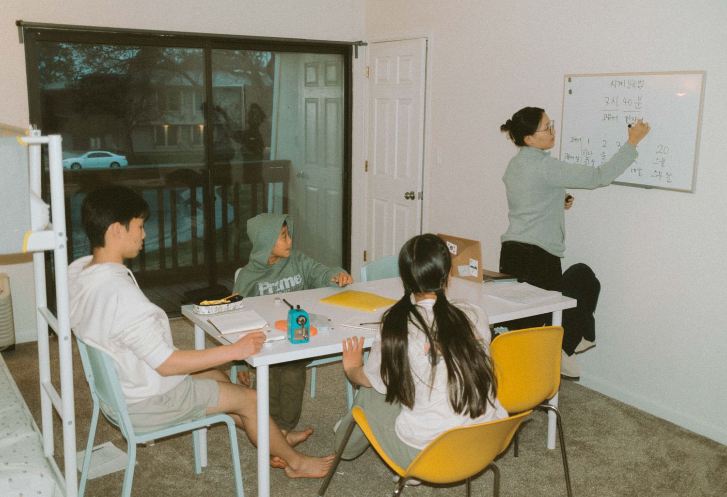 Students study at desks while a teacher writes on a whiteboard in a small classroom setting.