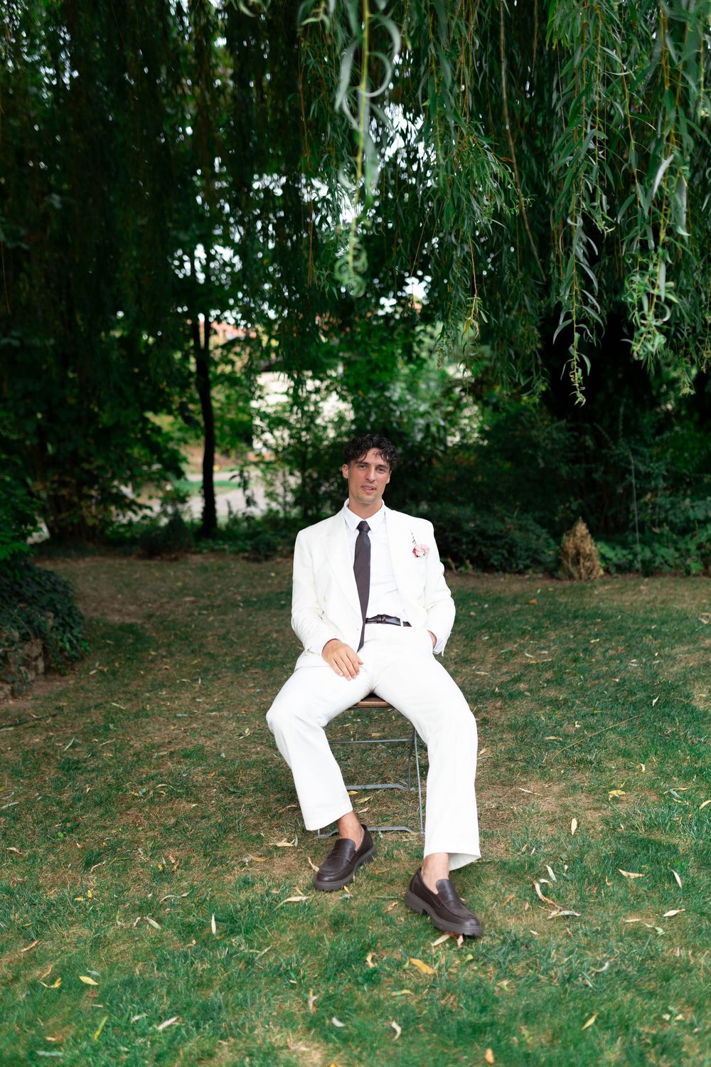 Young man in white suit and dark tie sitting on chair under weeping willow tree in lush green garden.