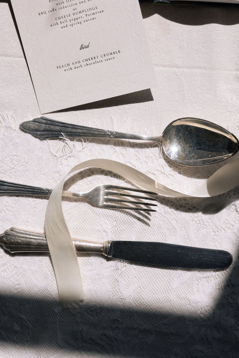 Silver cutlery set with fork, knife, and spoon tied with white ribbon on white linen tablecloth beside menu card.