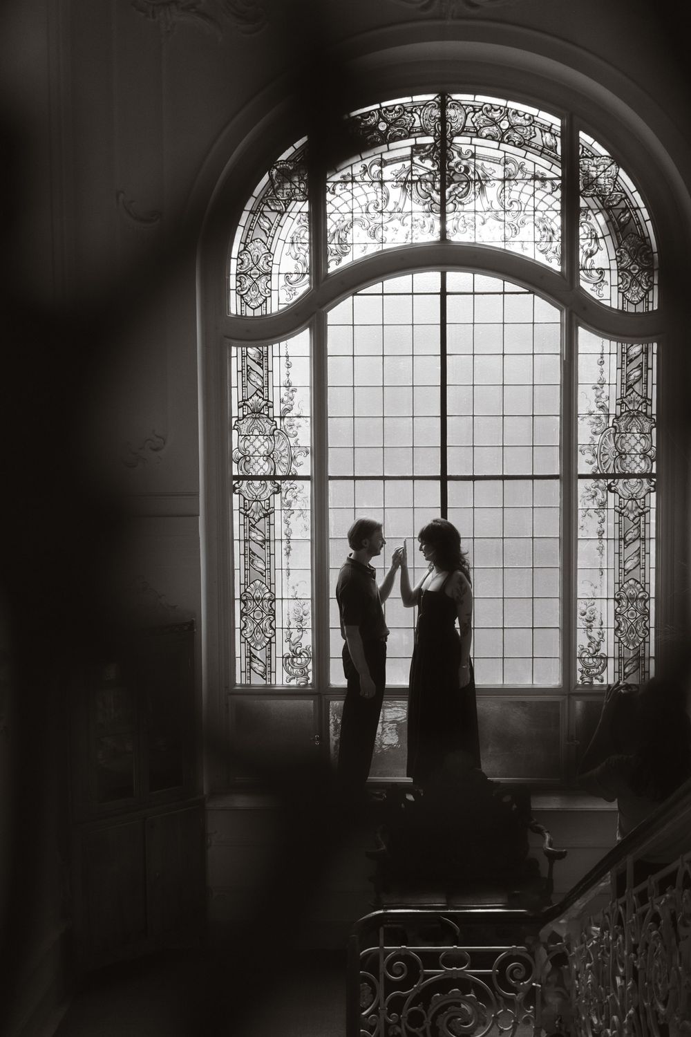 Black and white photo of a couple holding hands silhouetted against an ornate stained glass arched window.