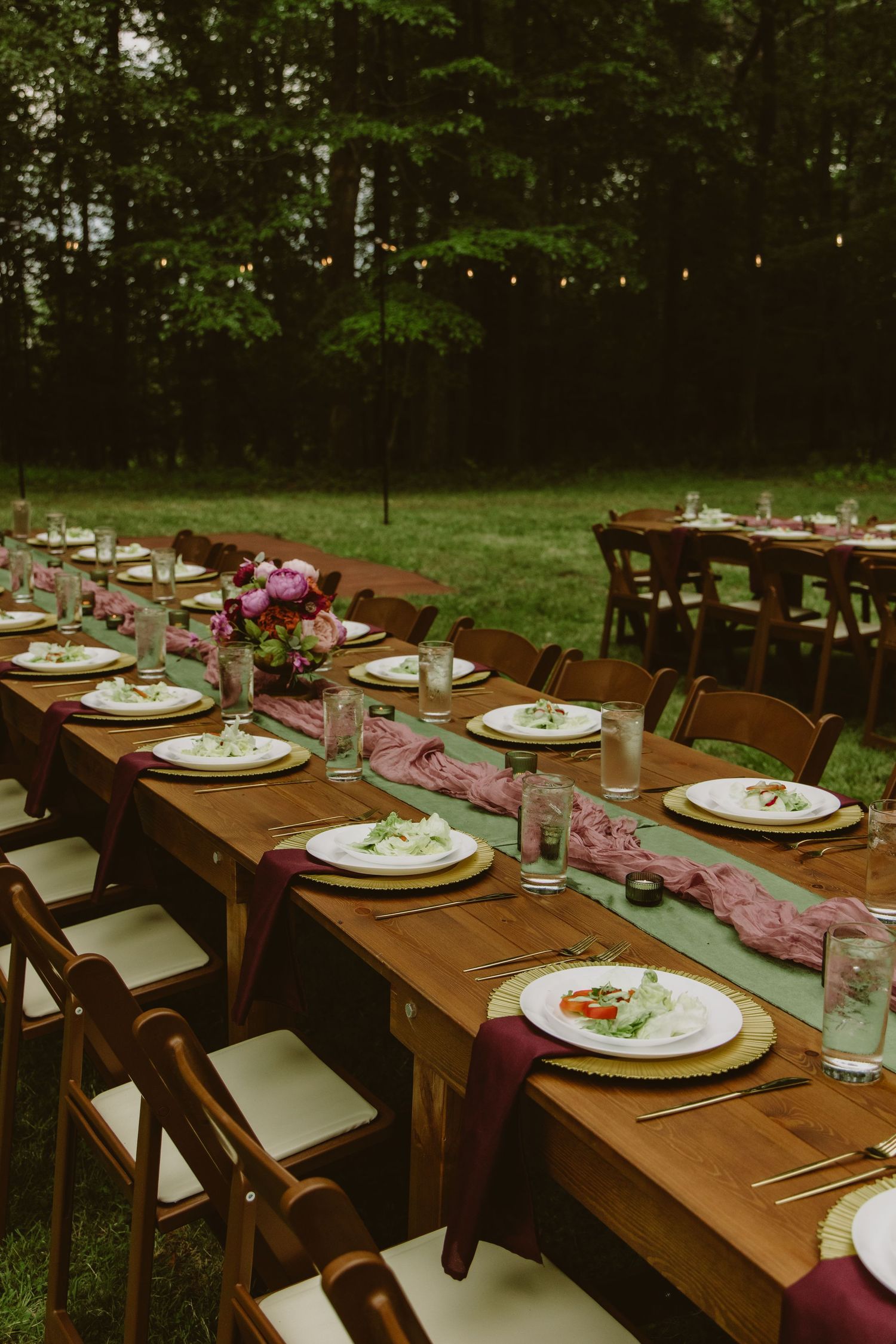 Elegant outdoor wedding table setting with candles, floral arrangements, white runner, and pink charger plates on wood table.