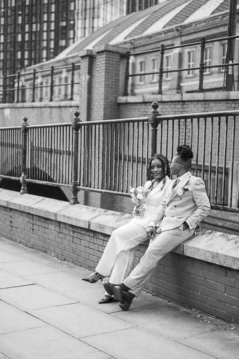 Black and white photo of a couple in wedding attire sitting on a brick wall with iron railings in an urban setting.