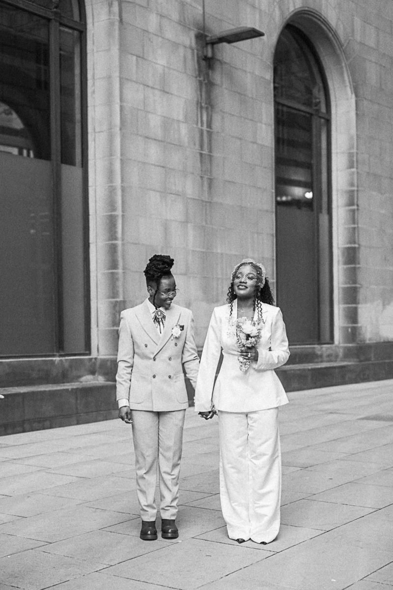 Two brides in white suits hold hands walking outside a stone building in a black and white wedding photo.