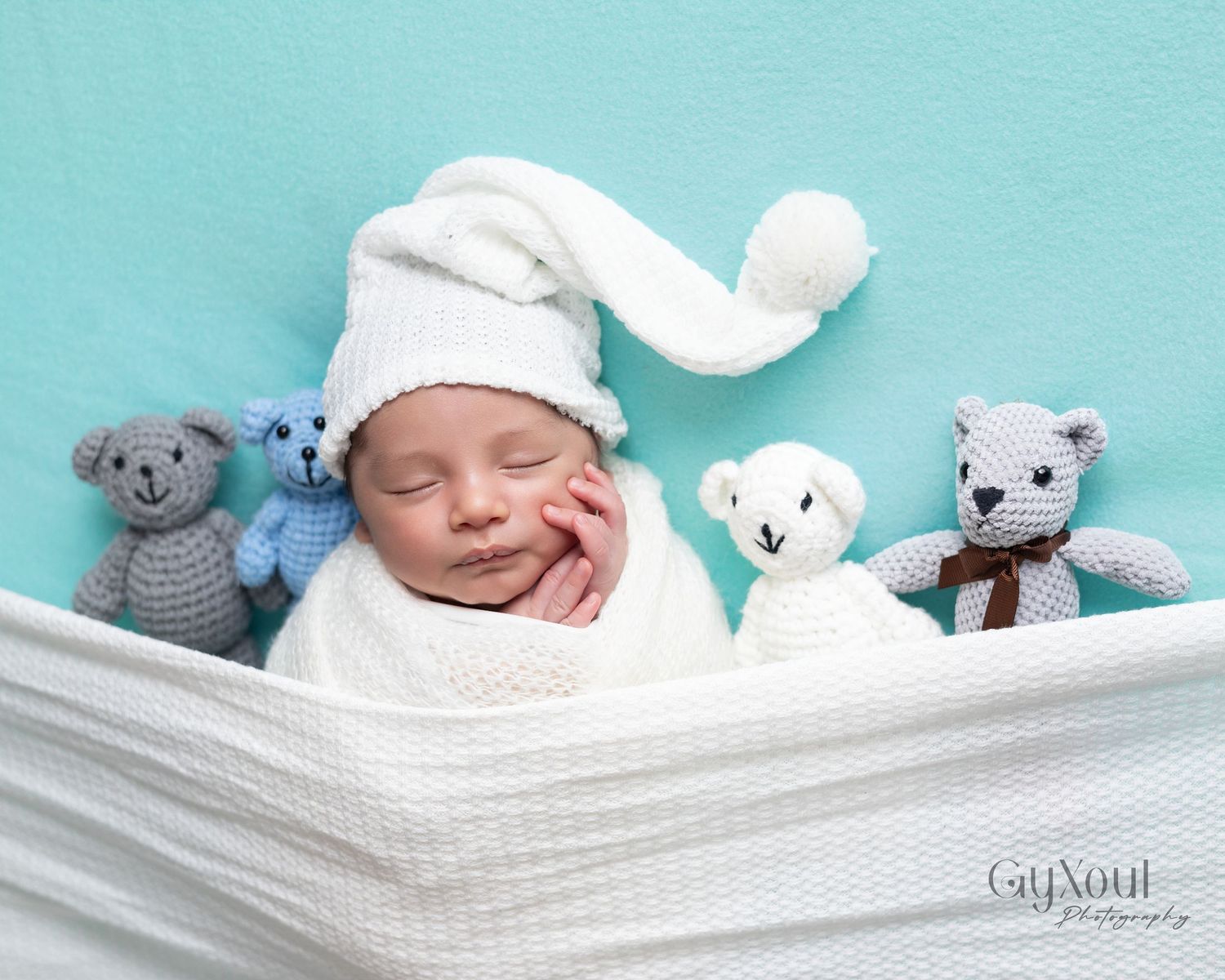 Newborn baby sleeping in white knit hat surrounded by four small crocheted teddy bears on teal background.