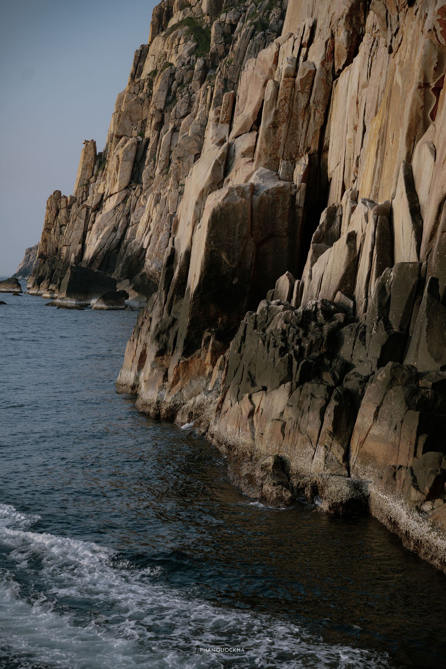 Dramatic rocky coastal cliffs rising from blue ocean waters, with wave foam at the base and rugged stone formations.