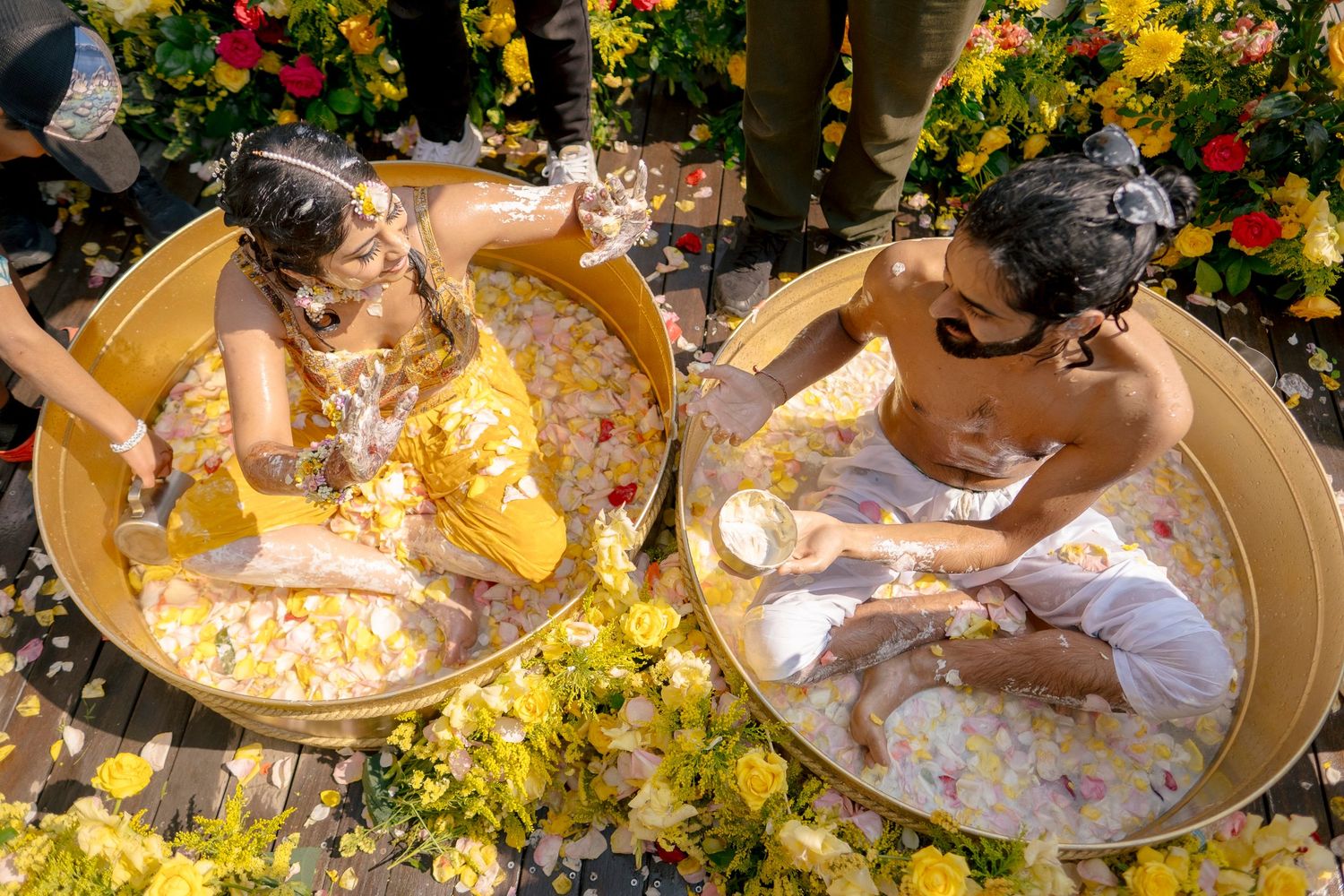 Two people bathing in golden tubs surrounded by yellow flowers and petals outdoors in a festive floral setting.