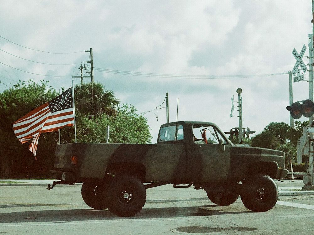 A lifted black pickup truck drives past an American flag and railroad crossing signs on a cloudy day.