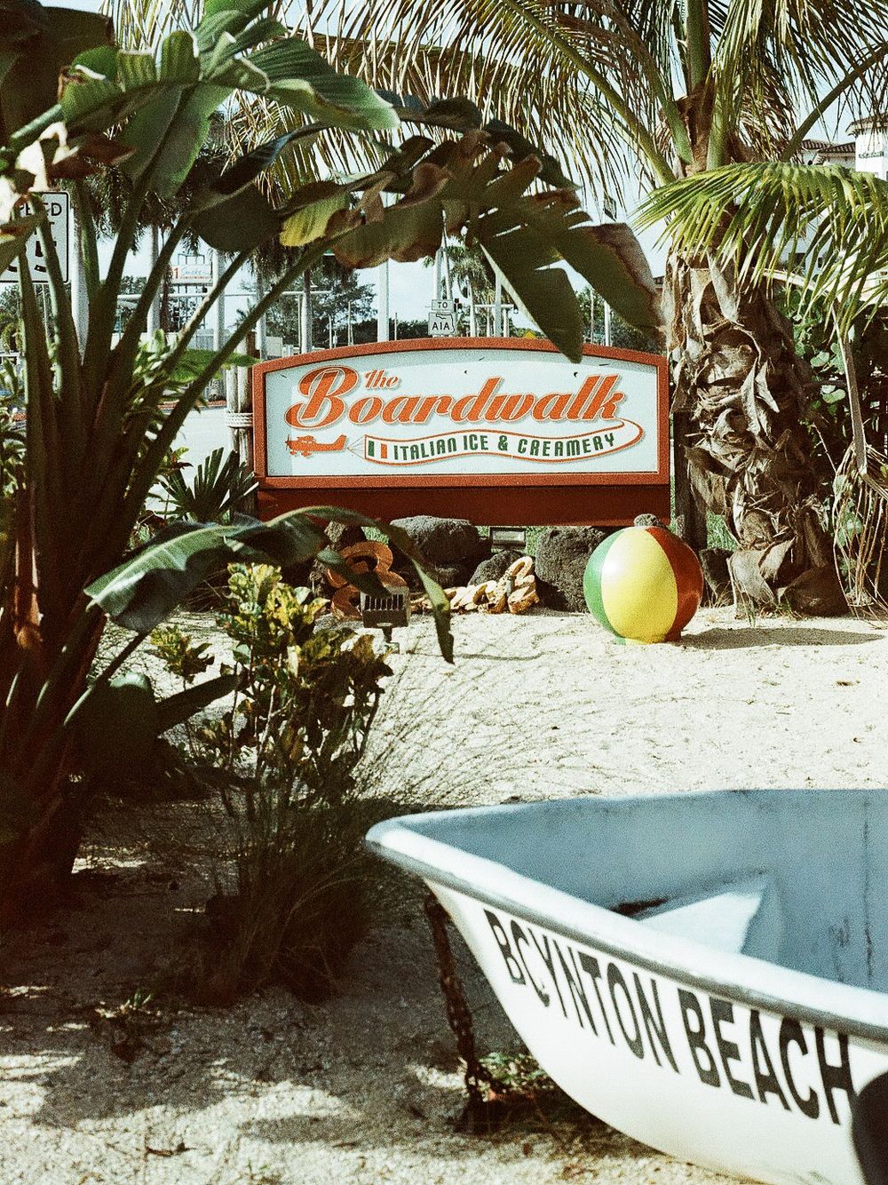 The Boardwalk Italian Ice and Creamery sign surrounded by tropical plants with a boat reading Boynton Beach in the foreground.