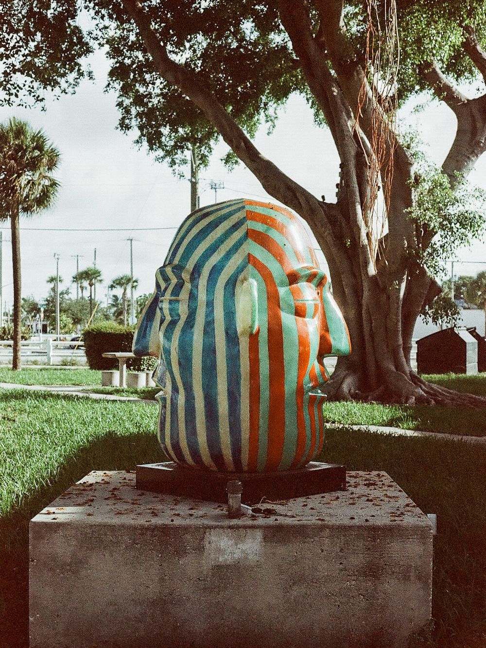 Colorful striped sculpture of a human head with blue and orange stripes displayed outdoors on a pedestal near trees.