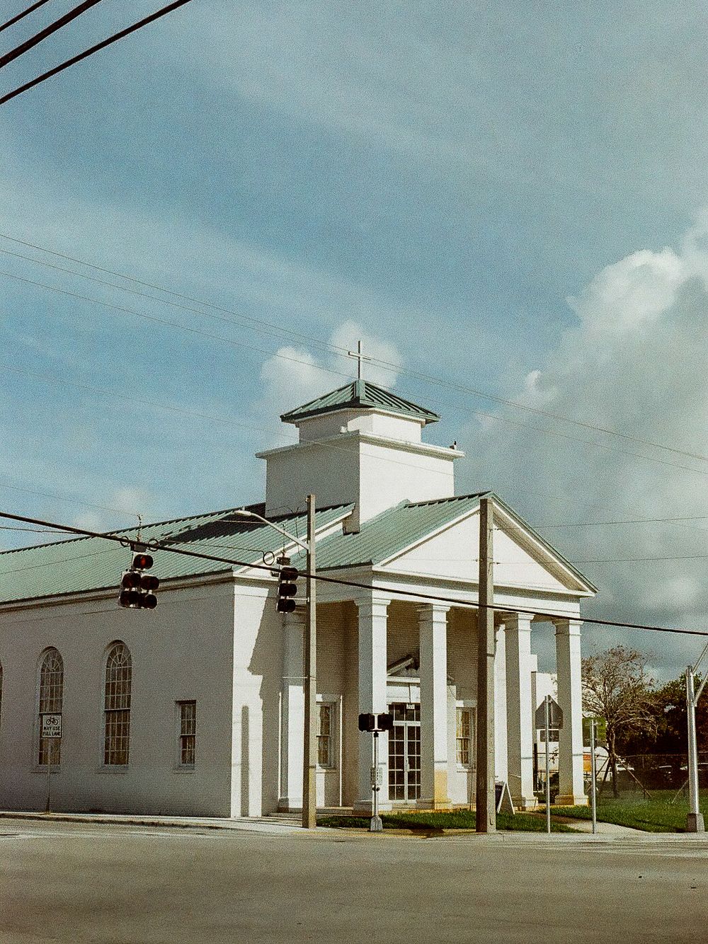 White church with green roof, columns, and cross on corner lot under blue sky.