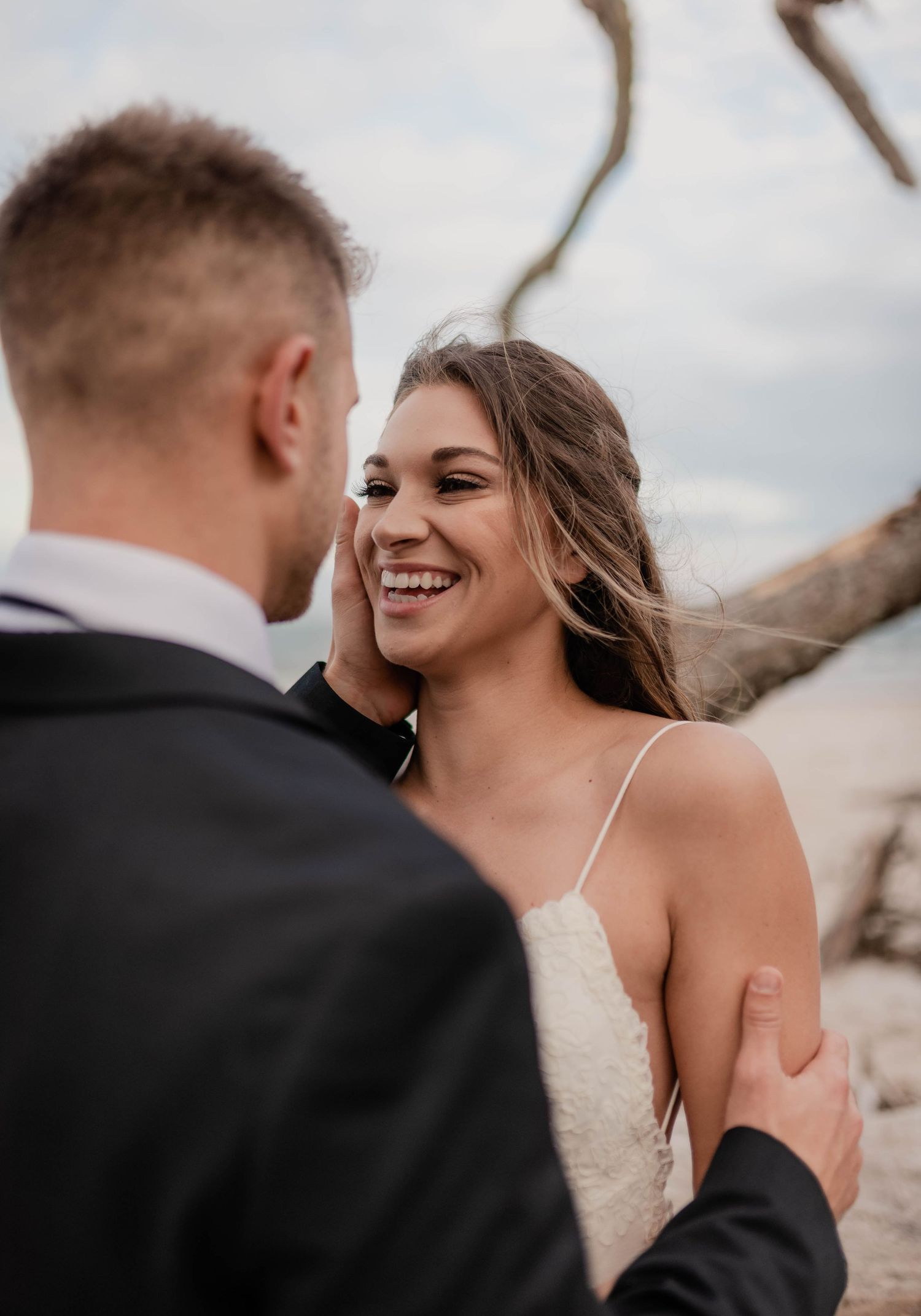 Bride in lace gown smiling joyfully at groom in black suit during outdoor beach wedding ceremony.