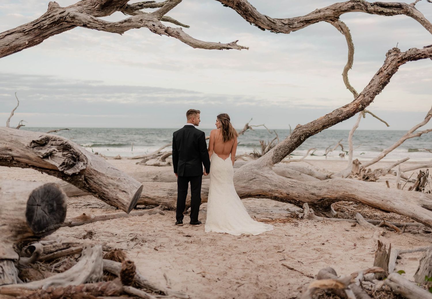 Bride and groom stand among driftwood on a sandy beach, gazing at the ocean on their wedding day.