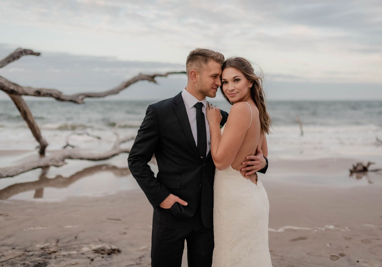Couple in wedding attire embracing on a beach with driftwood and ocean backdrop at dusk.